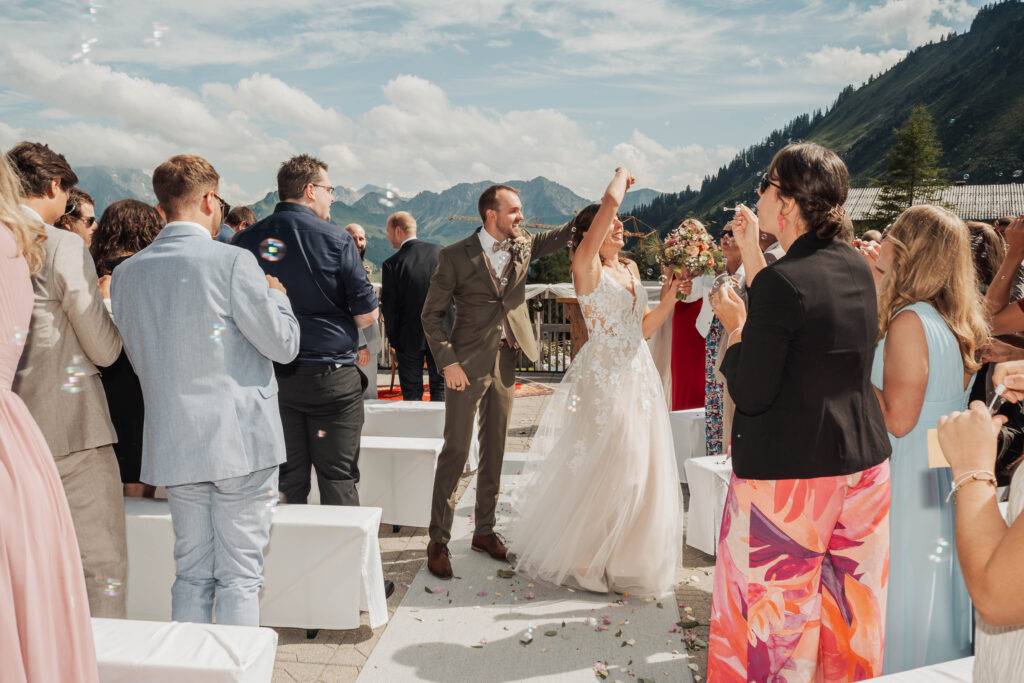 Gäste werfen Blüten oder Konfetti vor Bergkulisse.
