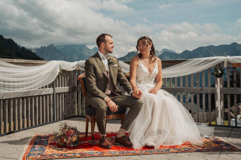 Braut und Bräutigam bei einer freien Trauung in den Bergen, sitzend und Händchen haltend, mit emotionalem Blick und alpiner Kulisse im Hintergrund.