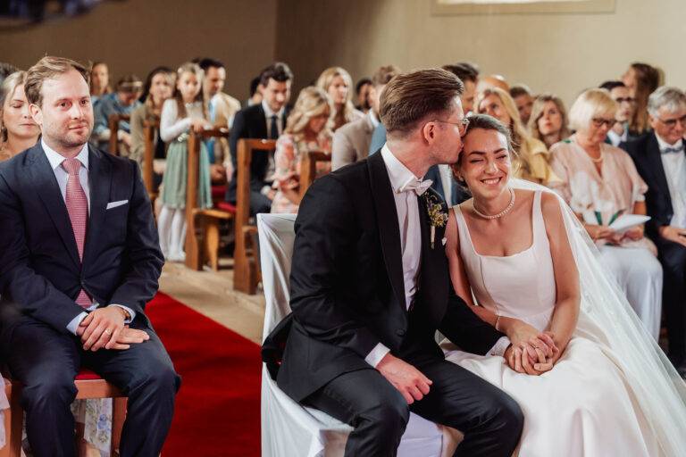 Braut und Bräutigam während der kirchlichen Trauung, liebevoller Kuss auf die Stirn mit Gästen im Hintergrund – emotionaler Moment für die Hochzeitsreportage.